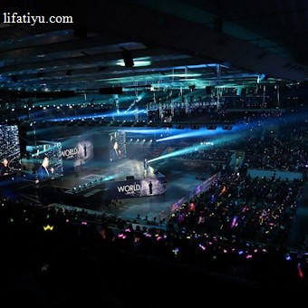 A large indoor arena filled with an audience watching a live performance. The stage features large screens displaying a performer and graphics, with bright spotlights and ambient lighting creating a dynamic atmosphere. The crowd is holding glow sticks, adding vibrant colors to the scene.