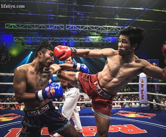 Two athletes are engaged in a dynamic kickboxing match inside a ring, surrounded by a cheering audience. One fighter, wearing red gloves and shorts, executes a high kick towards his opponent, who attempts to block the attack with his arm. The background features colorful lighting and promotional banners.