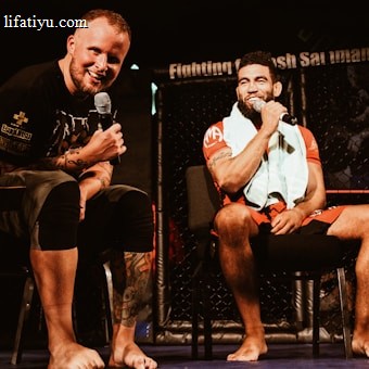 Two individuals are seated on a stage, each holding a microphone and smiling. They are in a sports-related setting with one wearing a casual black outfit and the other in sportswear, with a towel around his neck. The background includes elements of a fighting or martial arts cage.