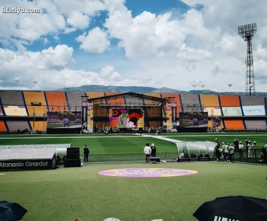 A sports stadium with a large stage set up on the field, hosting an outdoor event. Spectators wearing colorful hats sit in the foreground. The seating areas display vibrant colors, and a clear blue sky with scattered clouds is visible. Large screens display graphics related to the event.
