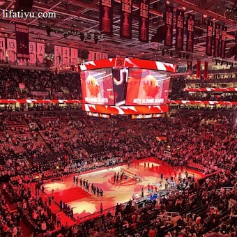 An indoor sports arena filled with spectators and a basketball court illuminated by red lighting. The scoreboards and video screens display promotional content, with numerous banners and championship flags hanging from the ceiling. Players and officials are gathered on the court, possibly in preparation for a game or event.
