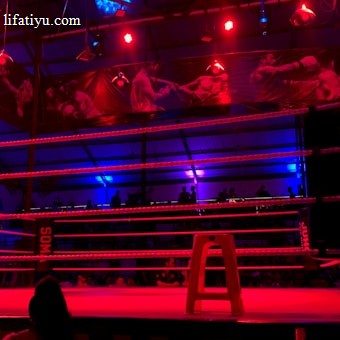 An empty boxing ring is illuminated by dramatic red and blue spotlights inside a dimly-lit arena. A small stool is placed in the center of the ring, surrounded by ropes. The arena is adorned with banners depicting action scenes, and a few silhouettes of spectators can be seen in the background.
